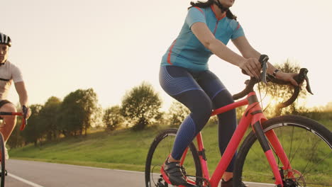 Dos-Ciclistas,-Un-Hombre-Y-Una-Mujer,-Viajan-Por-La-Carretera-En-Bicicletas-De-Carretera-Con-Cascos-Y-Ropa-Deportiva-Al-Atardecer-En-Cámara-Lenta